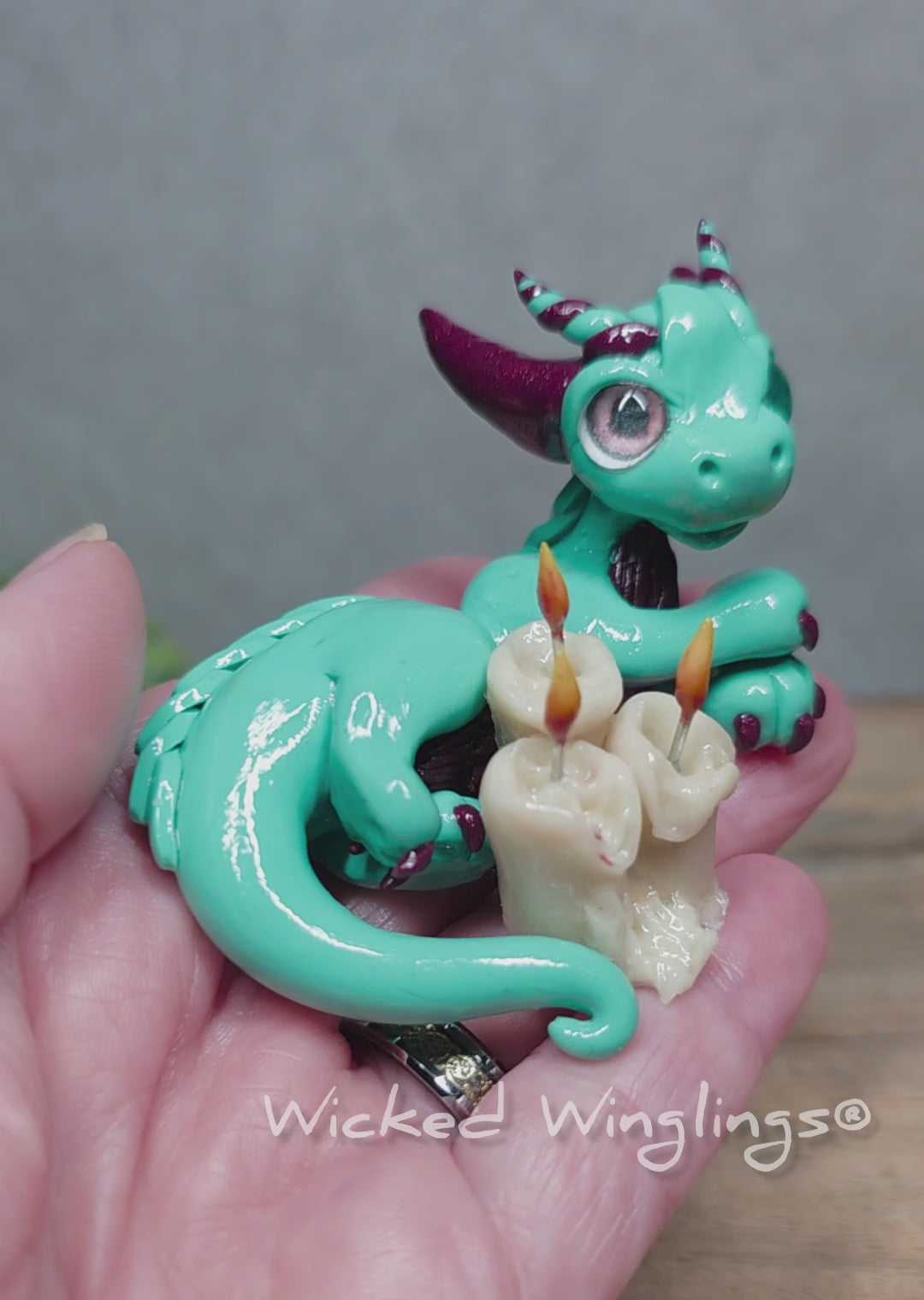 A video of a hand holding a clay dragon with candles against a wood surface with greenery. Text reads Wicked Winglings.