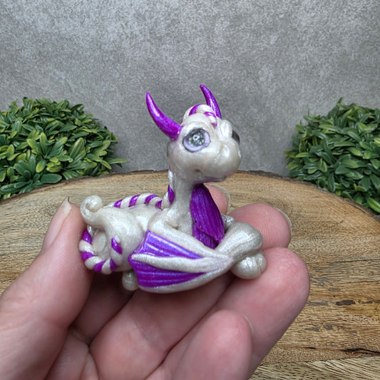 A hand holding a baby clay dragon against a wood background with greenery