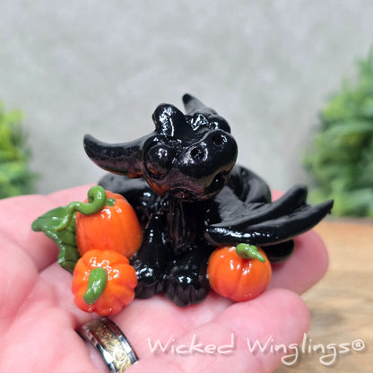 A hand holding a baby clay dragon sculpture with pumpkins against a wood background with greenery and text Wicked Winglings
