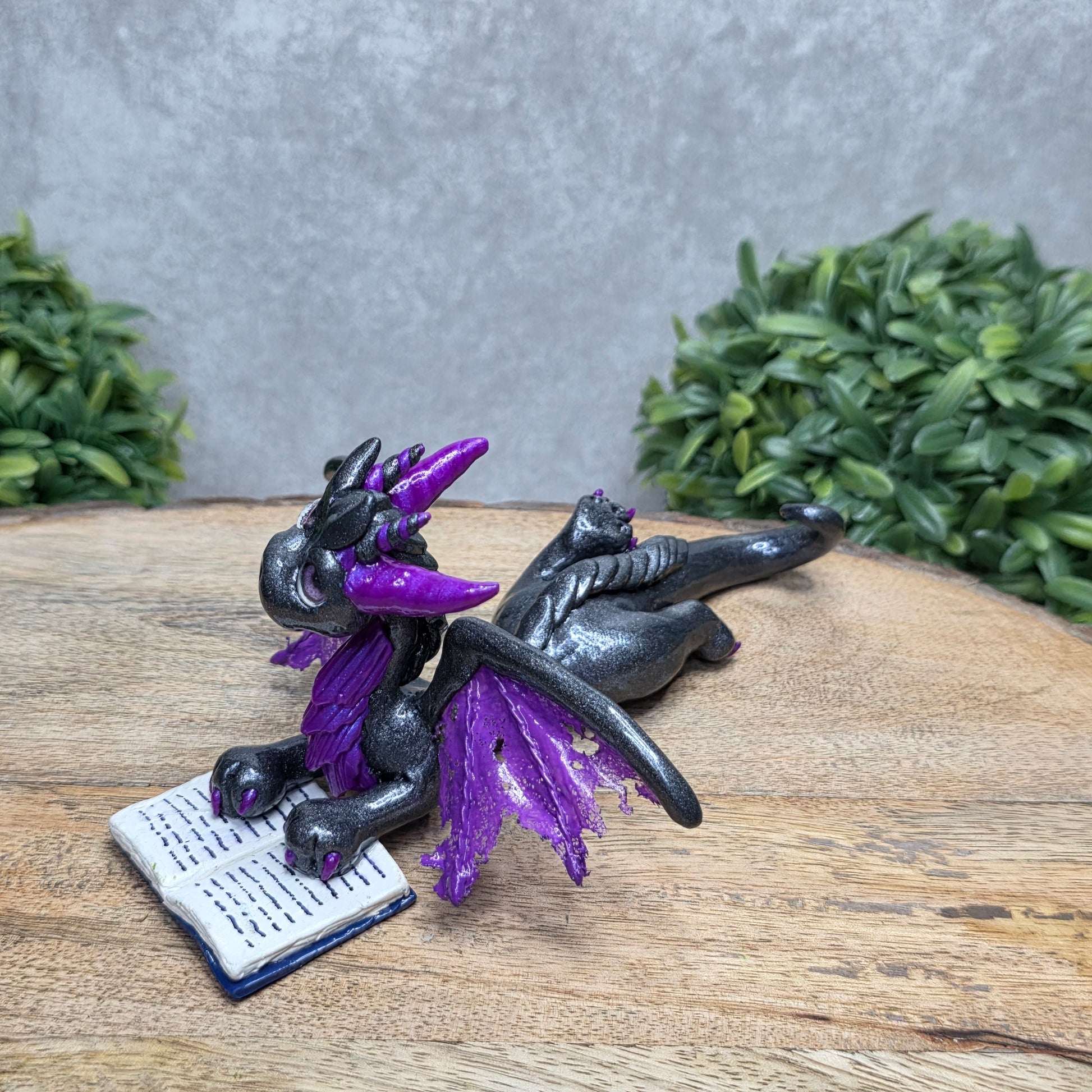 A dragon reading a book against a wood surface with greenery