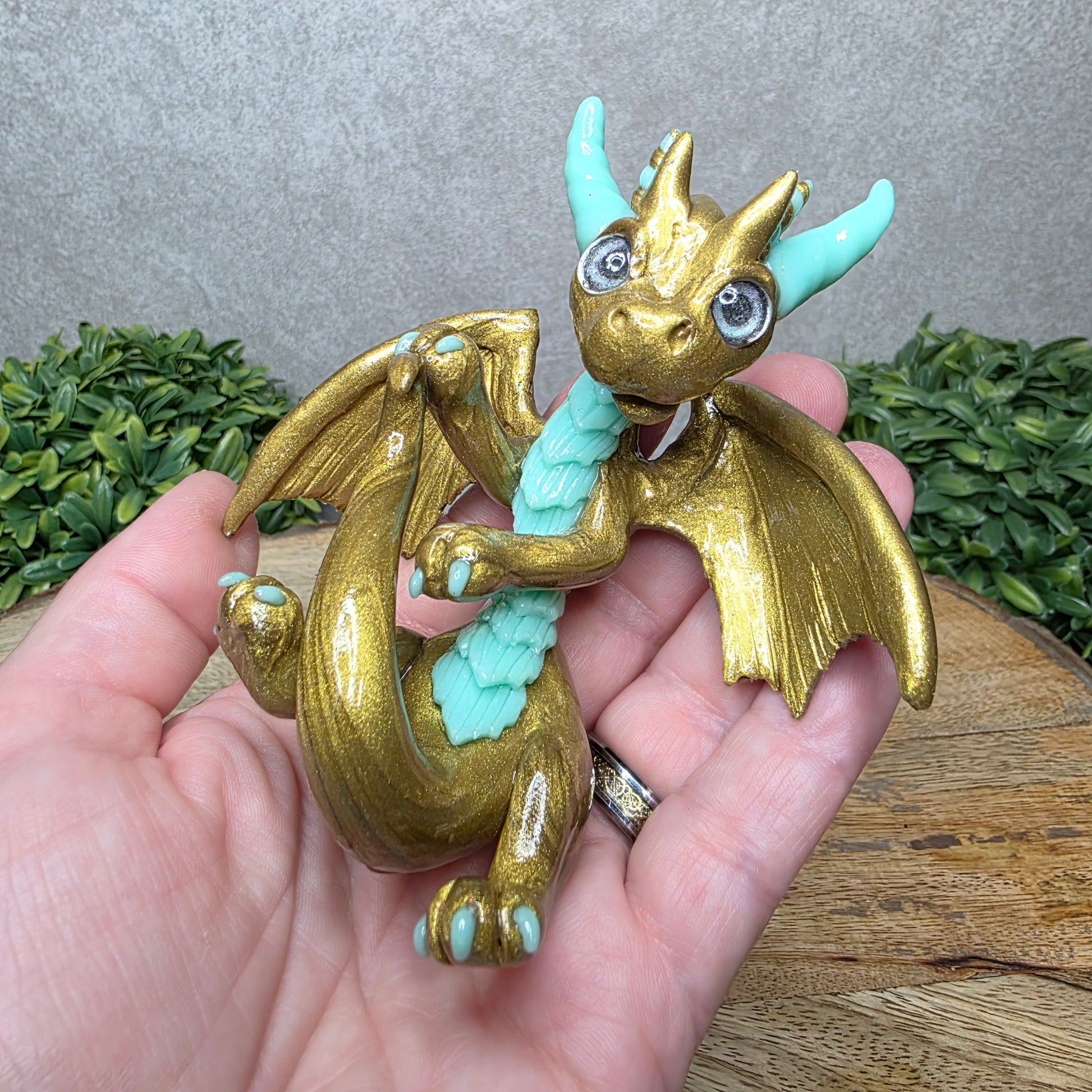 A hand holding a clay dragon against a wood background with greenery