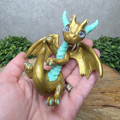 A hand holding a clay dragon against a wood background with greenery