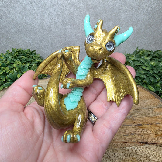 A hand holding a clay dragon against a wood background with greenery