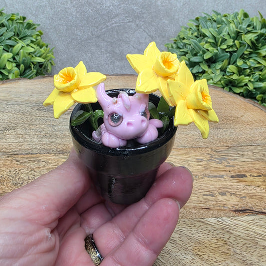 Hand holding a clay baby dragon in a pot with daffodils against a wood surface with greenery.