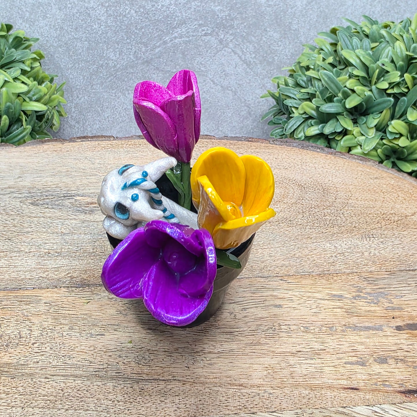 Side view of a baby clay dragon in a pot with tulips against a wood surface with greenery.