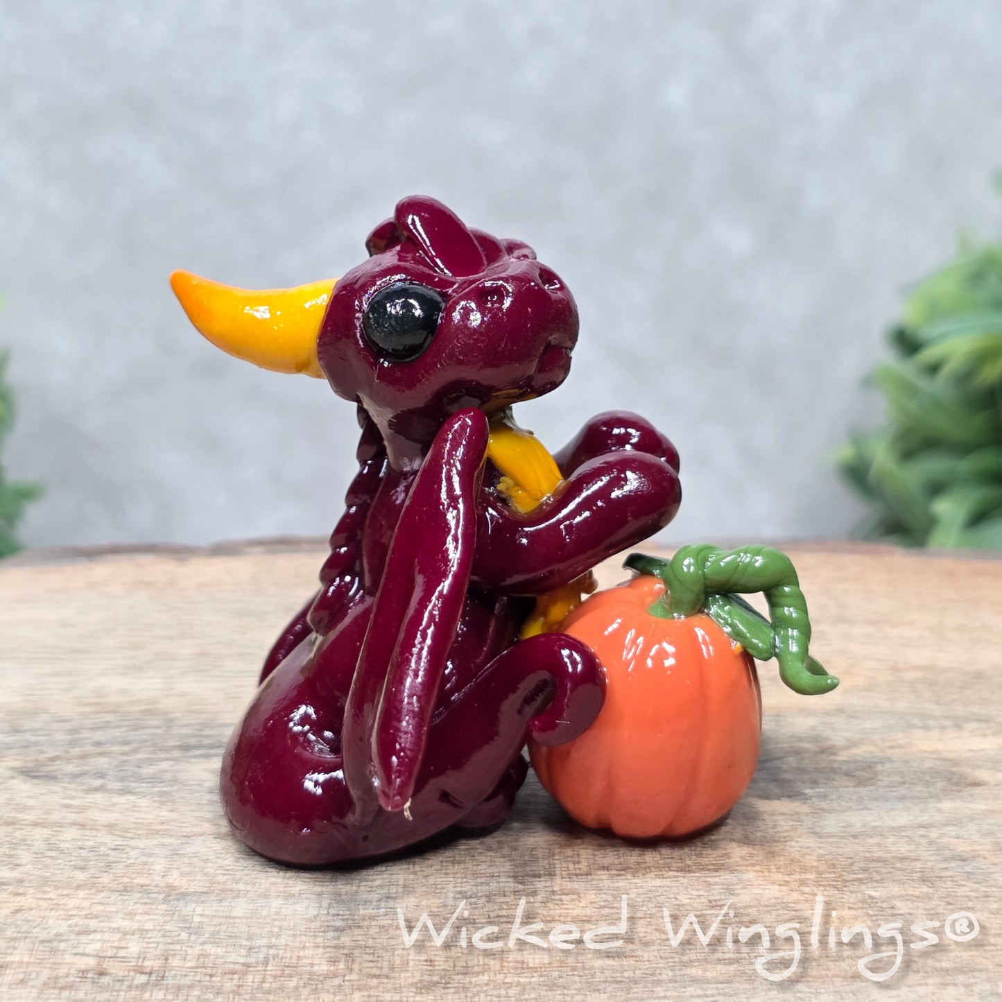 A baby clay dragon sculpture with a pumpkin against a wood background with greenery and text Wicked Winglings