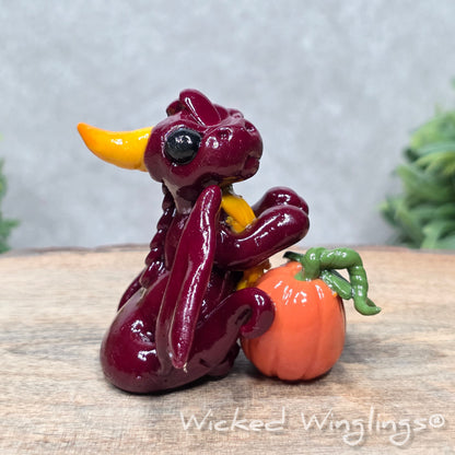 A baby clay dragon sculpture with a pumpkin against a wood background with greenery and text Wicked Winglings
