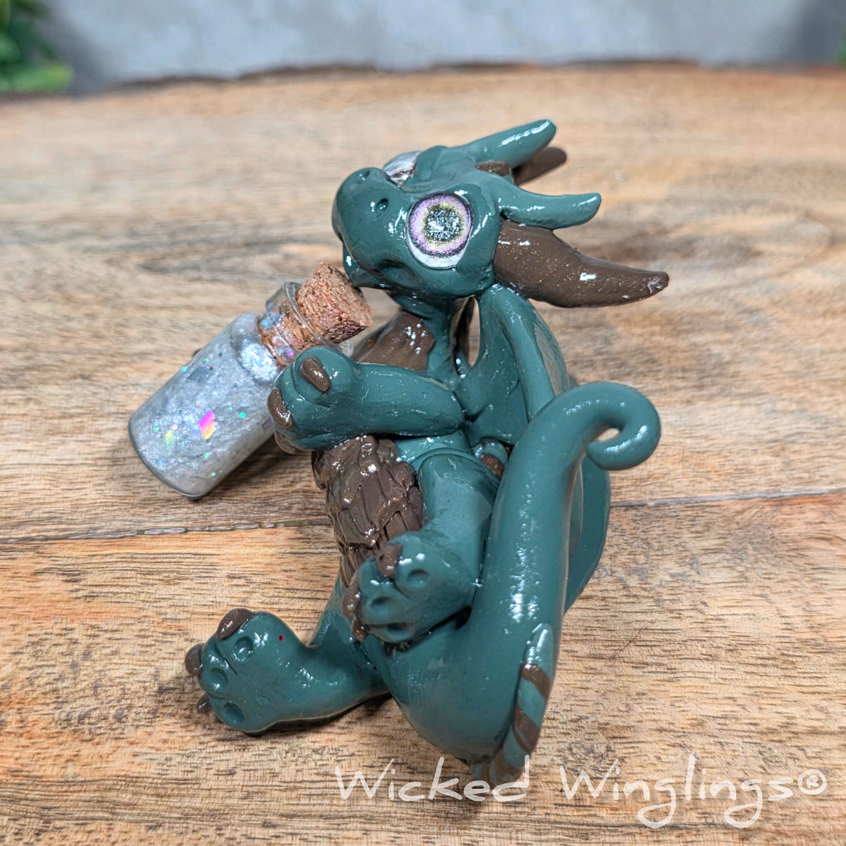 A clay dragon with a potion bottle on a wood surface. Text reads Wicked Winglings.
