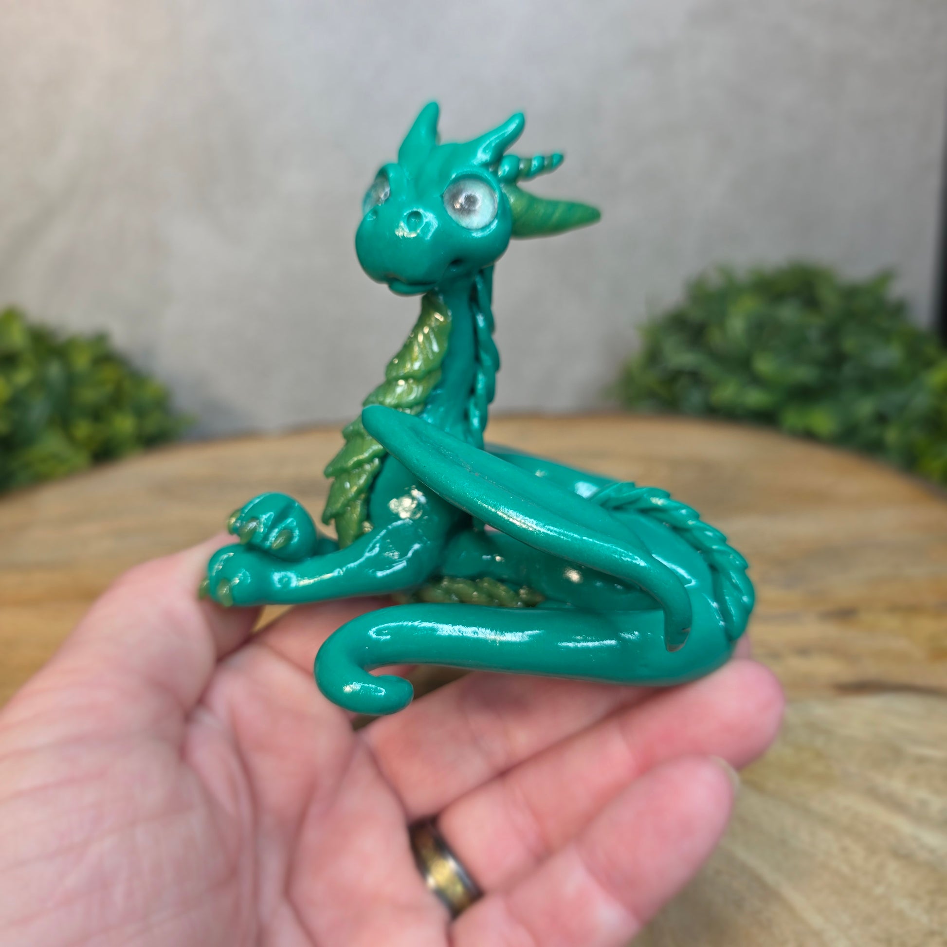 A hand holding a green clay dragon against a blurred wood surface
