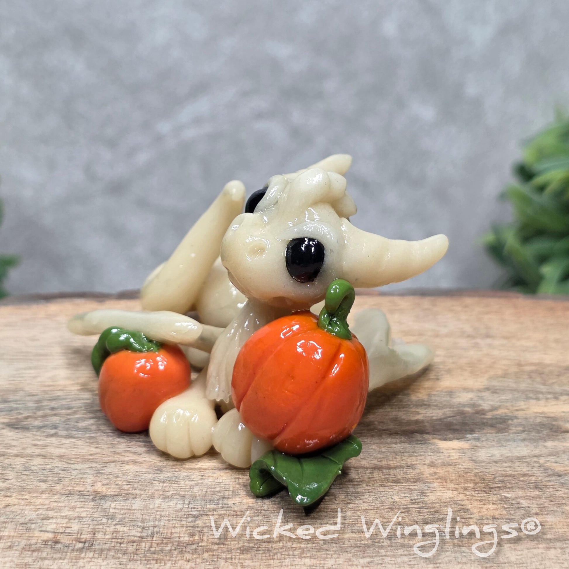 A baby clay dragon sculpture with pumpkins against a wood background with greenery and text Wicked Winglings