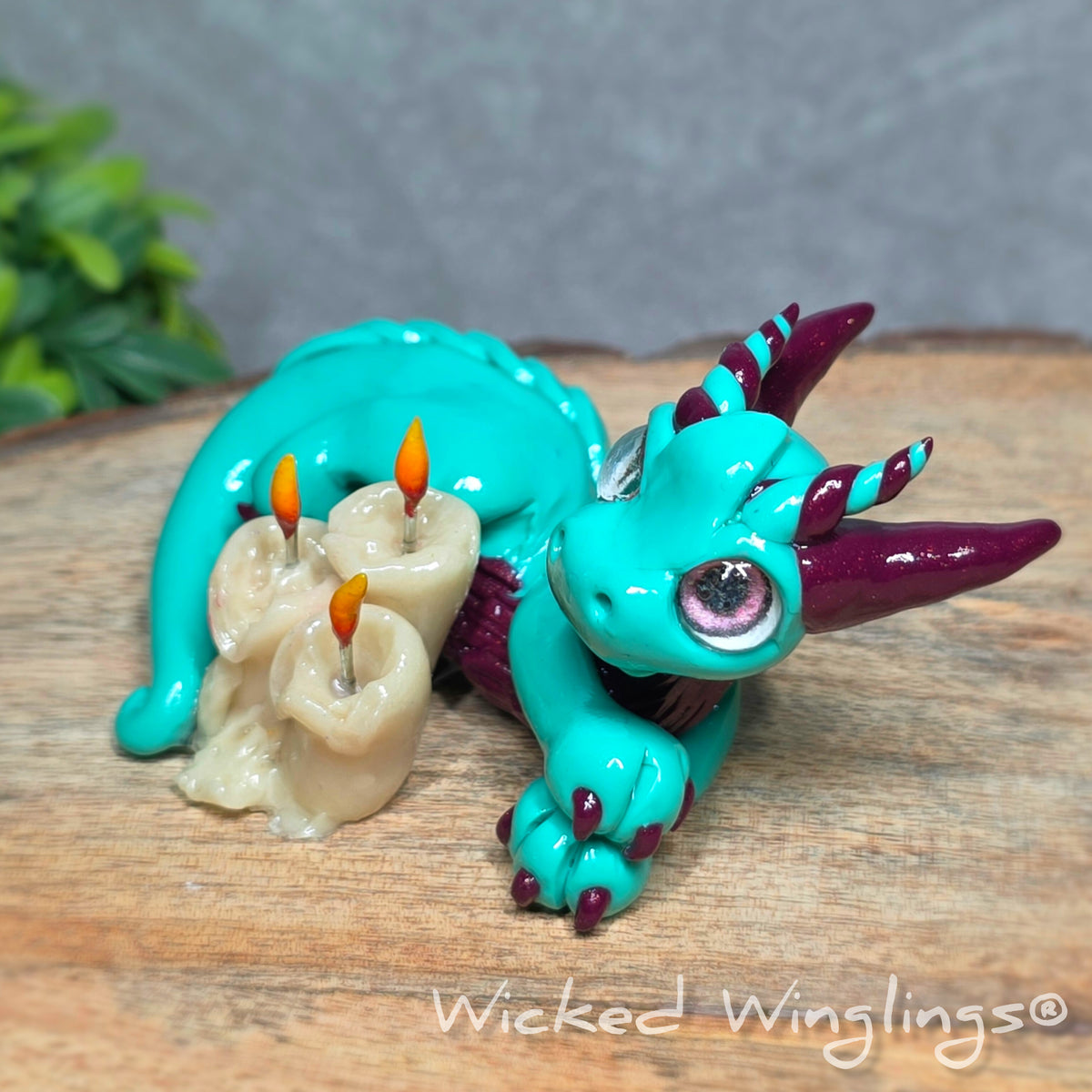 Side view of a clay dragon with candles against a wood surface with greenery. Text reads Wicked Winglings.