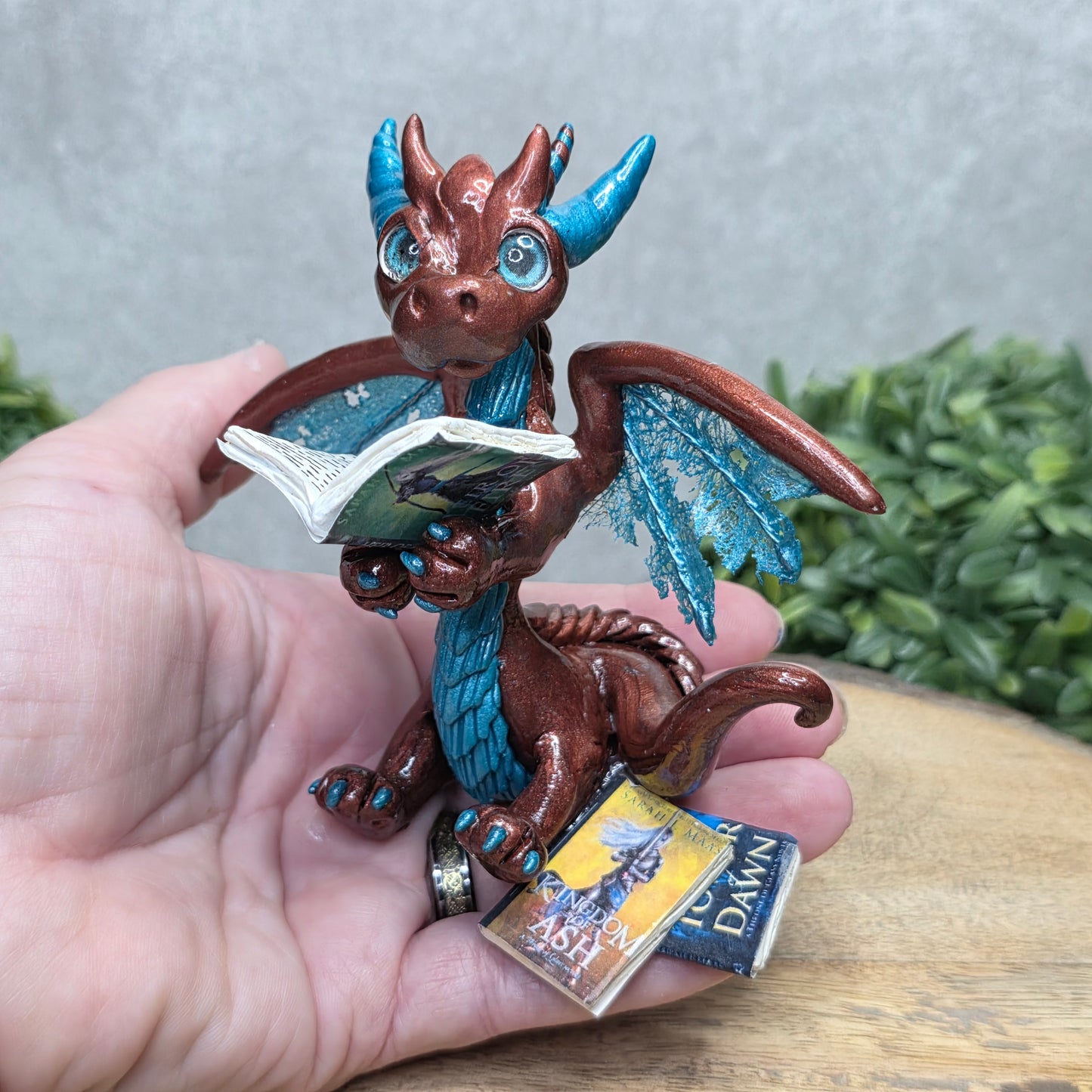 Hand holding a dragon reading a book against a wood surface with greenery