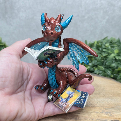 Hand holding a dragon reading a book against a wood surface with greenery