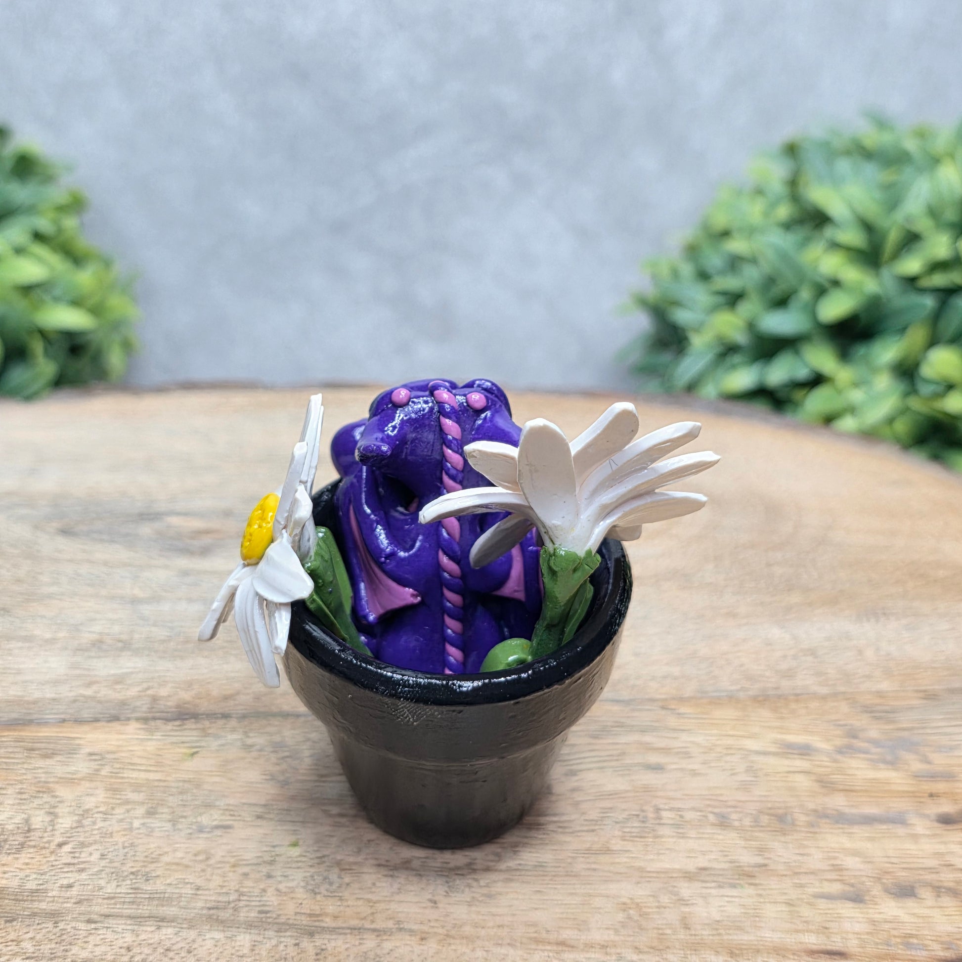 Back view of a clay baby dragon in a pot with daisies against a wood surface with greenery.