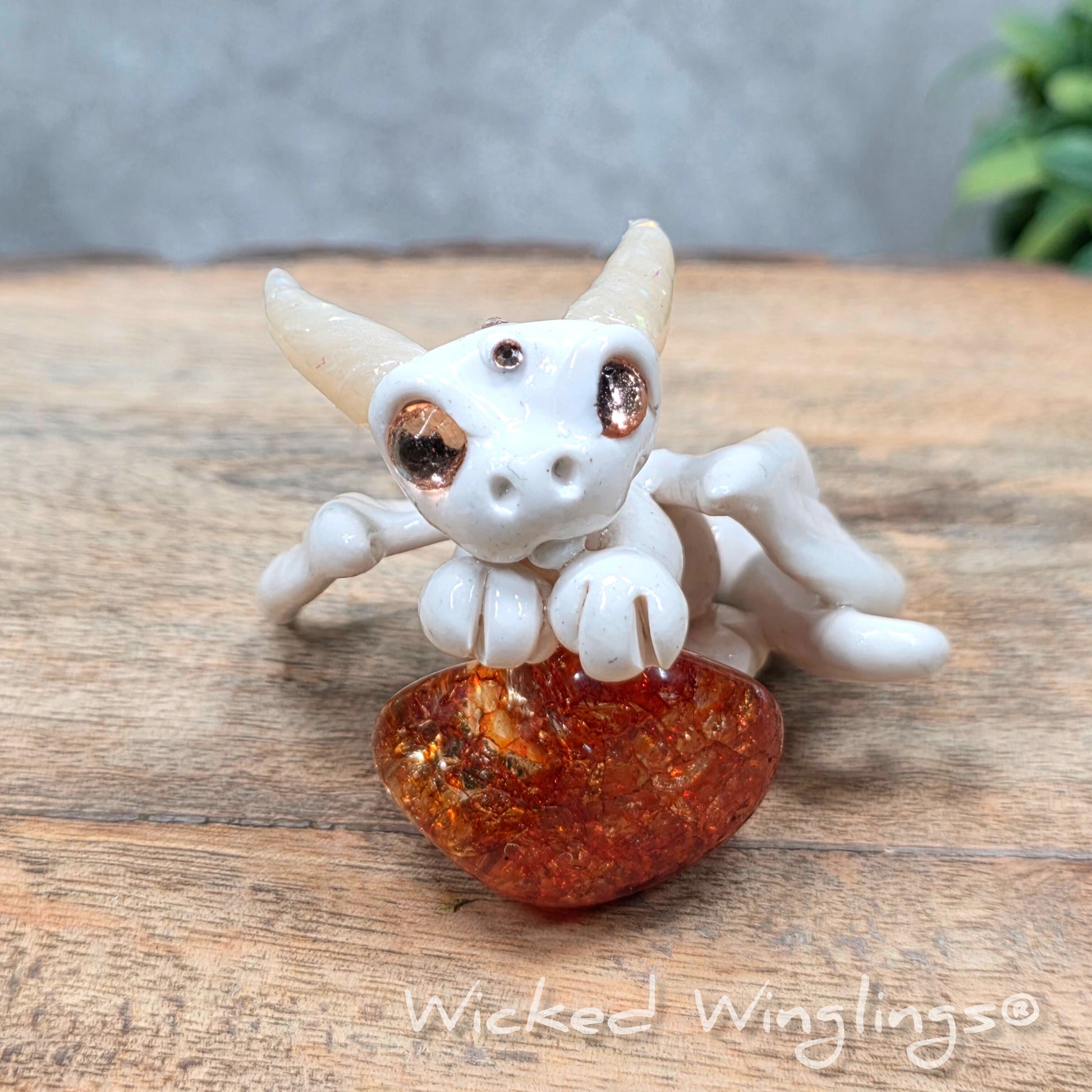 A baby clay dragon sculpture with a crackled quartz against a wood background with greenery and text Wicked Winglings