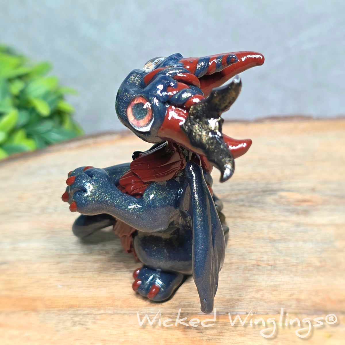 Side view of a clay dragon with bats sitting on a wood surface with greenery. Text reads Wicked Winglings.