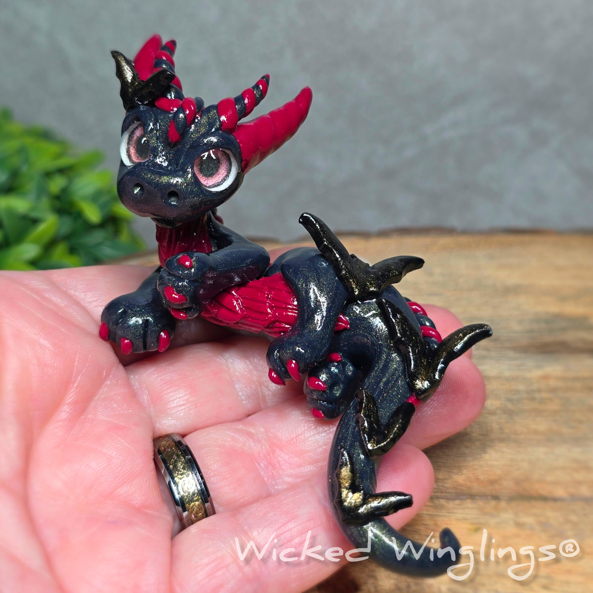 A hand holding a clay dragon with bats against a wood surface with greenery. Text reads Wicked Winglings.
