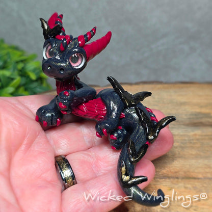 A hand holding a clay dragon with bats against a wood surface with greenery. Text reads Wicked Winglings.