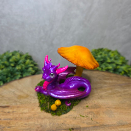 Purple dragon figurine with a yellow mushroom on a wooden surface with greenery