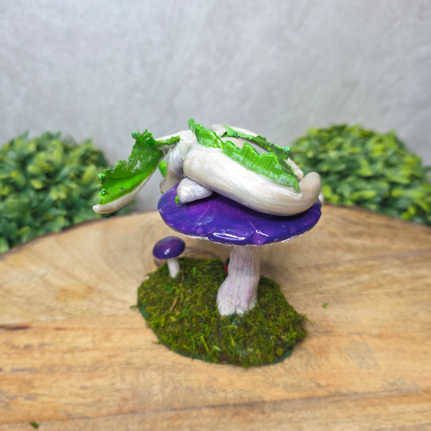 A clay dragon figurine on a  mushroom on a wooden surface with greenery.