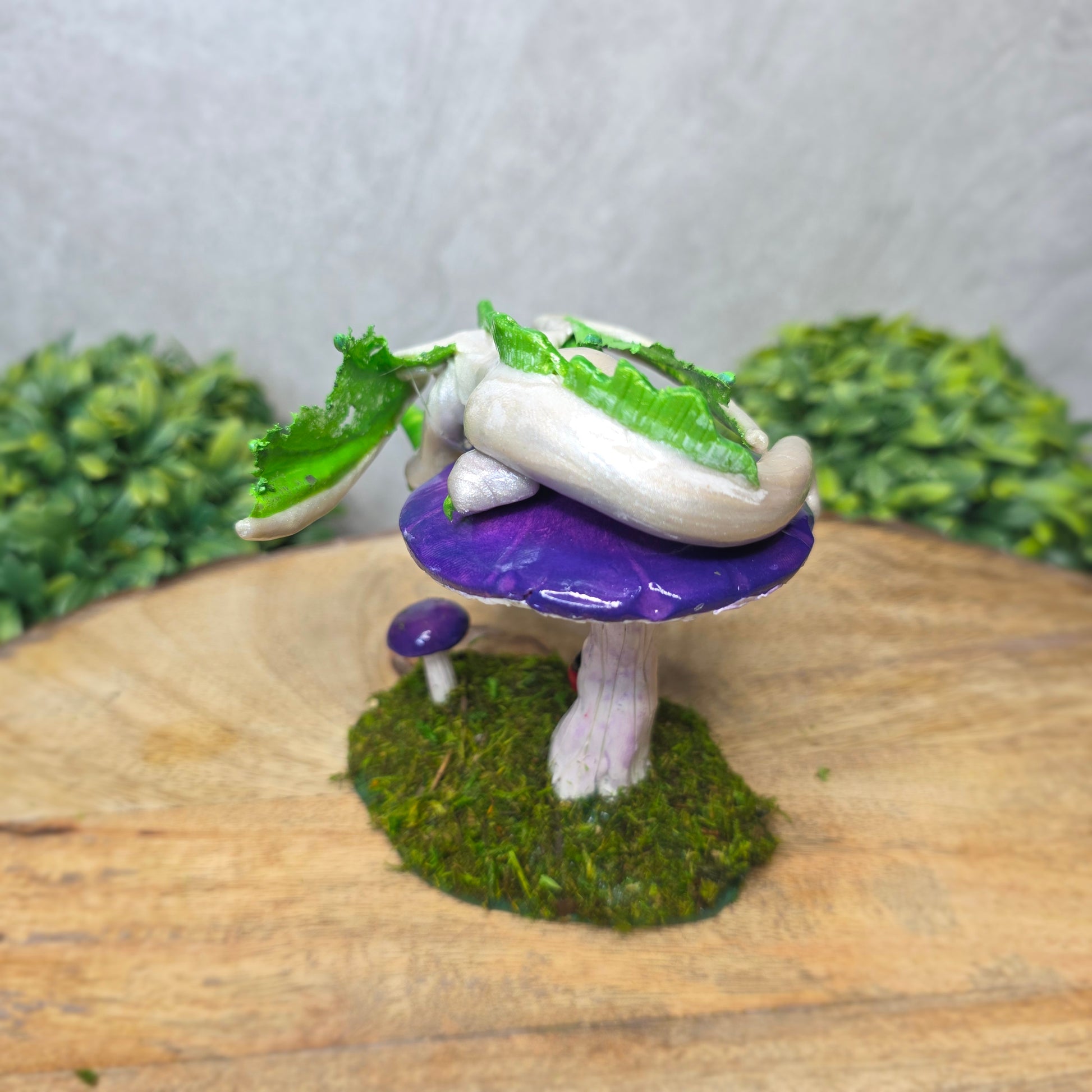 A clay dragon figurine on a  mushroom on a wooden surface with greenery.