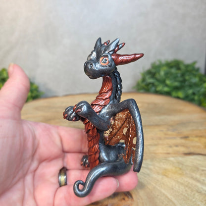 A hand holding a graphite and bronze clay dragon against a blurred wood surface