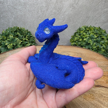A hand holding a flocked clay dragon sculpture on a wood surface with greenery.