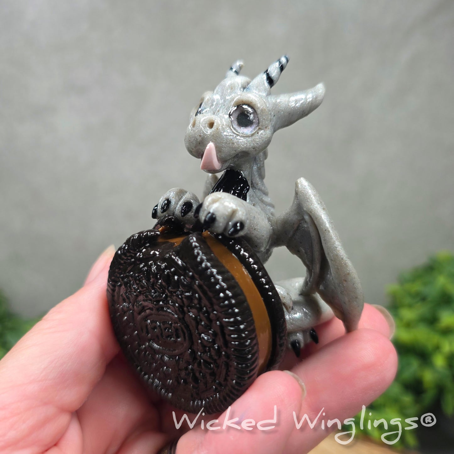 A hand holding a clay dragon with a cookie with a Wicked Winglings® watermark.