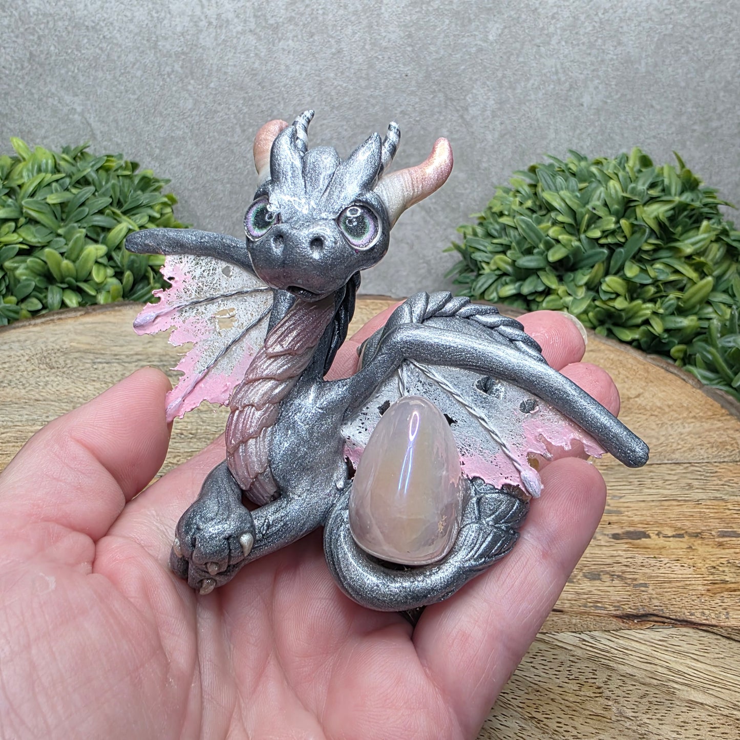 Hand holding a clay dragon with a rose quartz crystal against a wood surface with greenery.