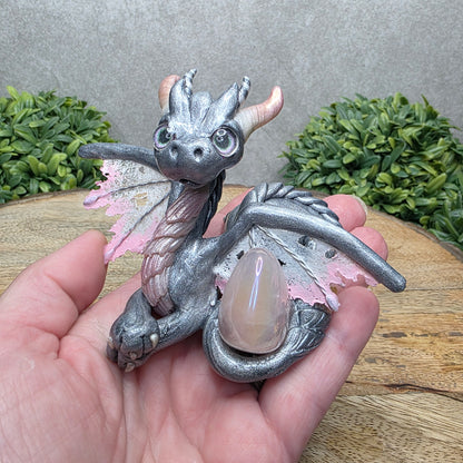 Hand holding a clay dragon with a rose quartz crystal against a wood surface with greenery.