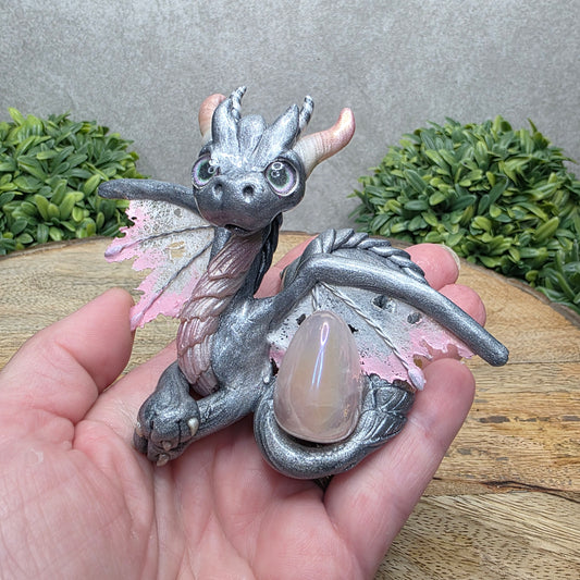 Hand holding a clay dragon with a rose quartz crystal against a wood surface with greenery.