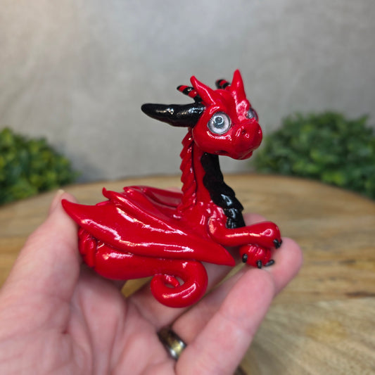 A hand holding a red and black clay dragon against a blurred wood surface