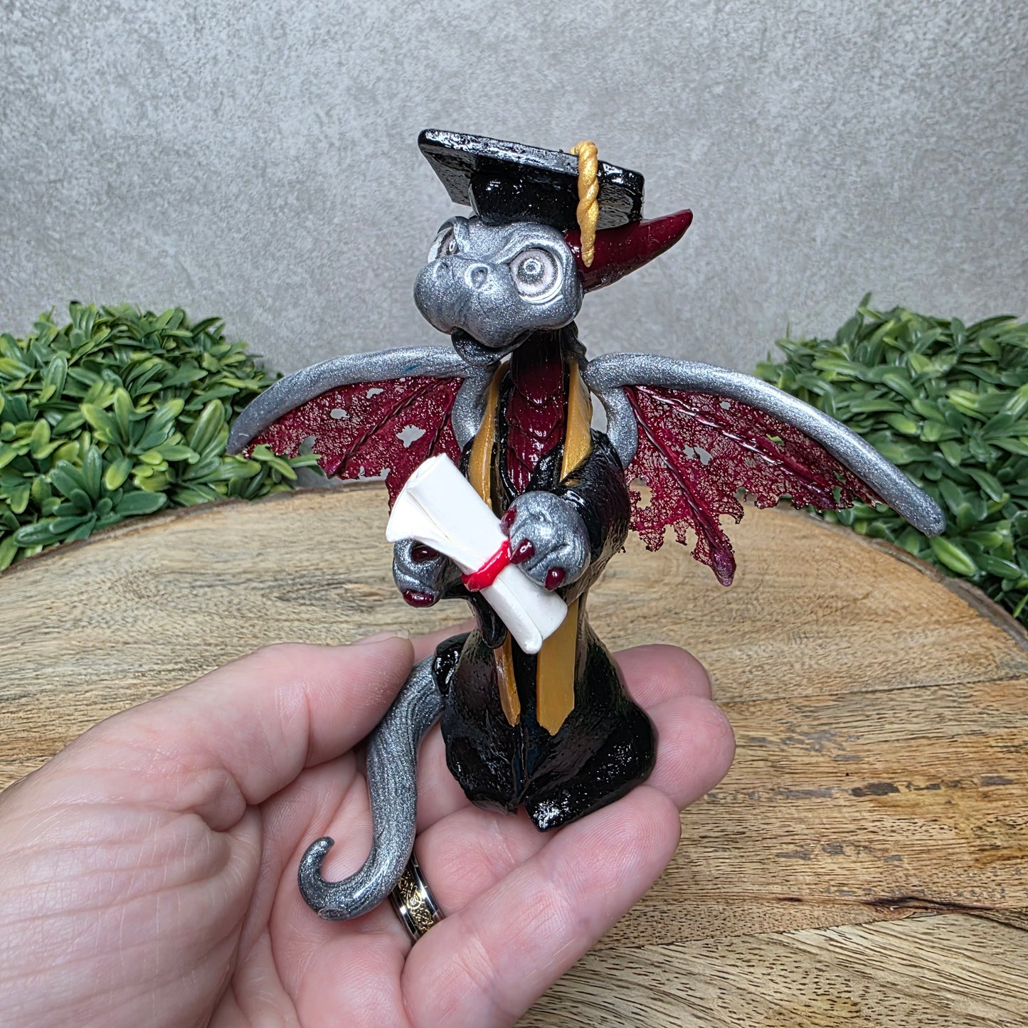 A hand holding a clay dragon in a graduation cap and gown holding a diploma