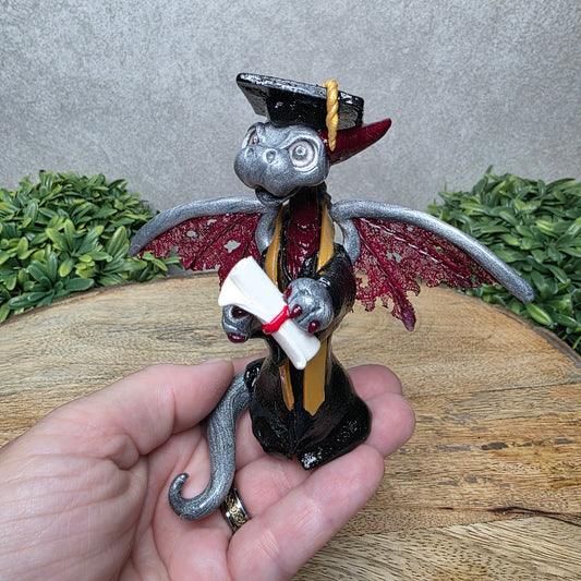 A hand holding a clay dragon in a graduation cap and gown holding a diploma