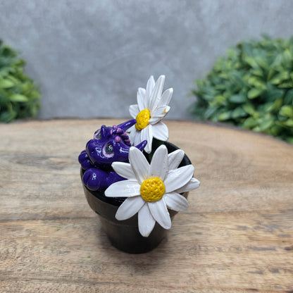 Side view of a clay baby dragon in a pot with daisies against a wood surface with greenery.