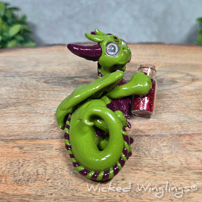 A clay dragon with a potion bottle on a wood surface. Text reads Wicked Winglings.