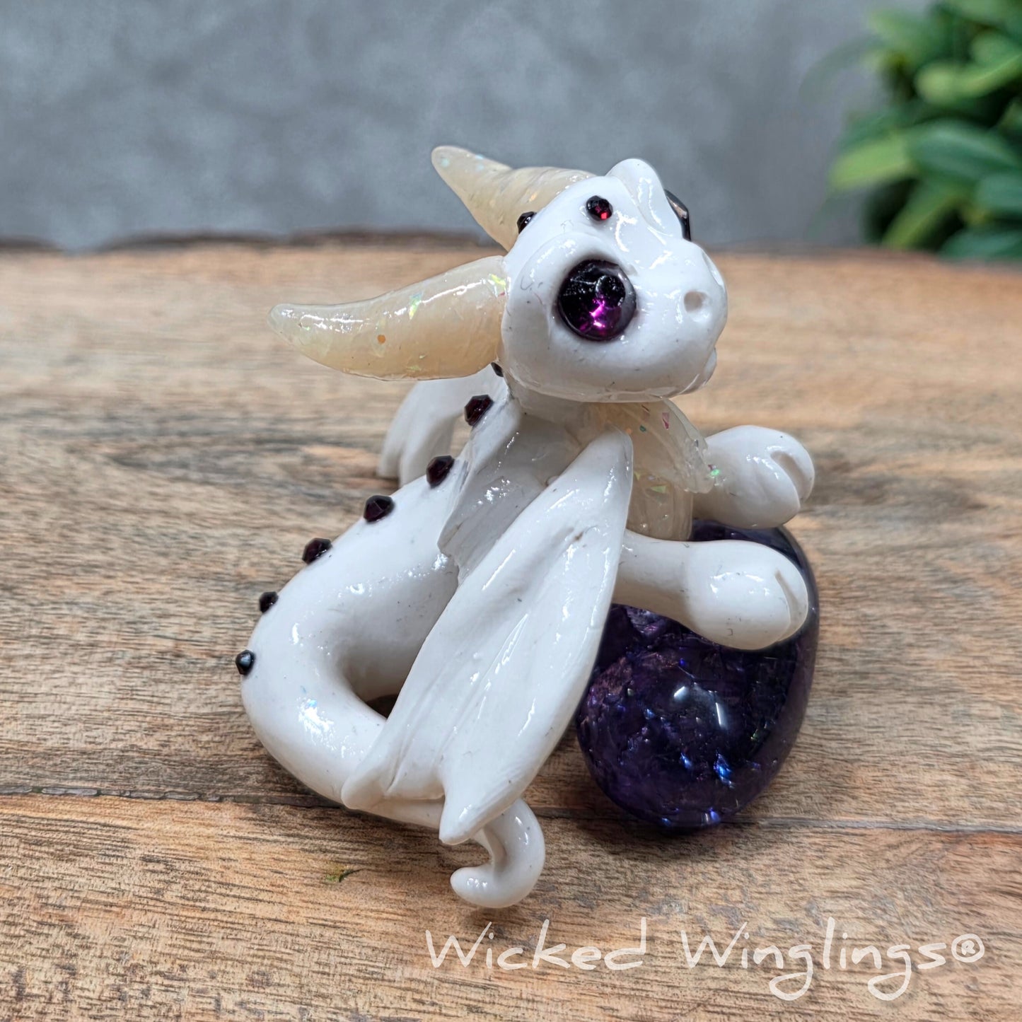 A baby clay dragon sculpture with a crackled quartz against a wood background with greenery and text Wicked Winglings