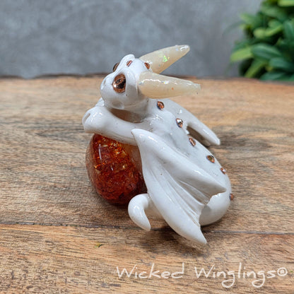 A baby clay dragon sculpture with a crackled quartz against a wood background with greenery and text Wicked Winglings