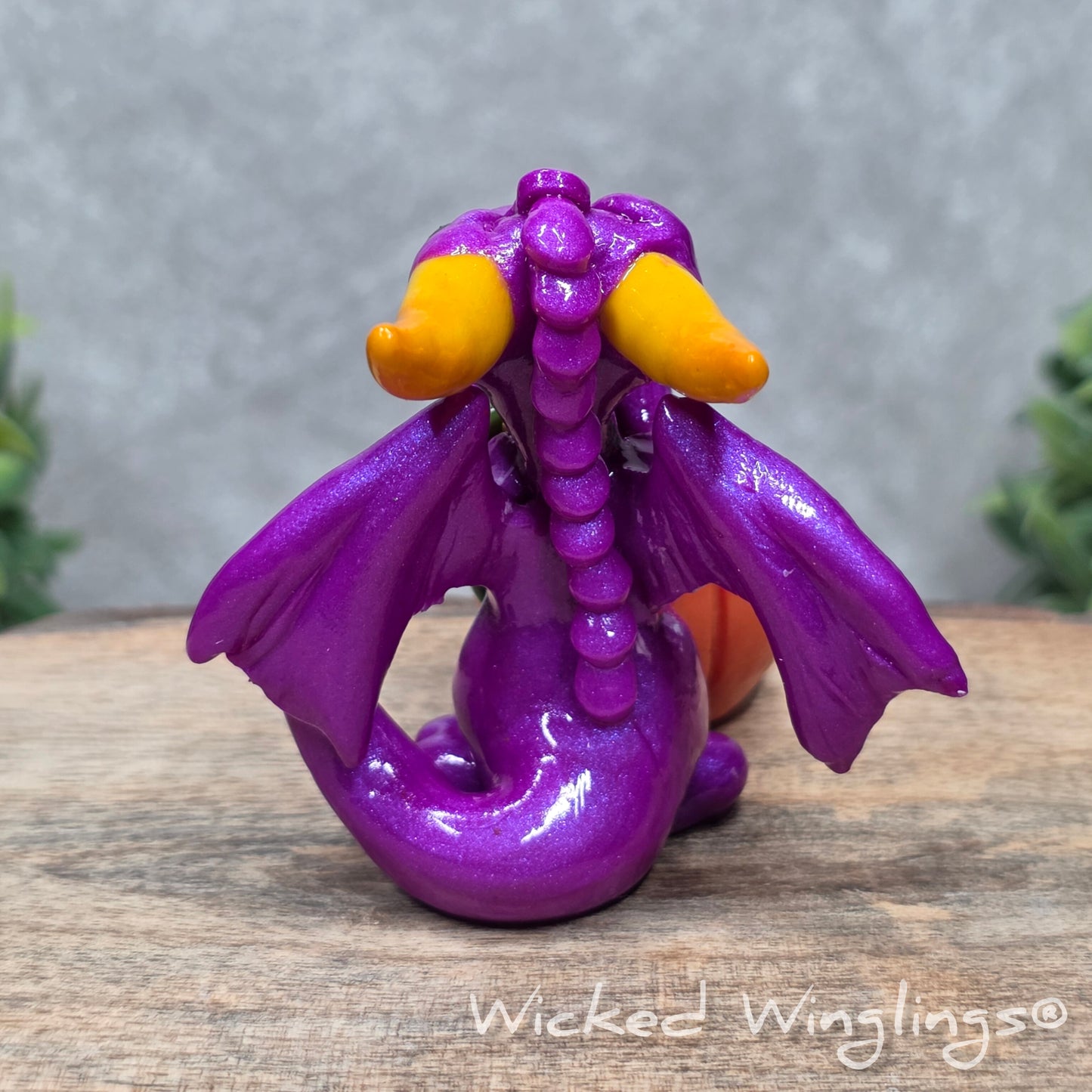 A baby clay dragon sculpture with a pumpkin against a wood background with greenery and text Wicked Winglings