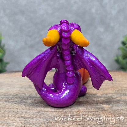 A baby clay dragon sculpture with a pumpkin against a wood background with greenery and text Wicked Winglings
