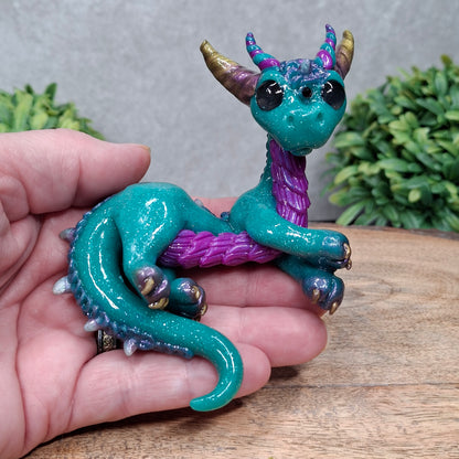  A hand holding a clay dragon against a wood background with greenery