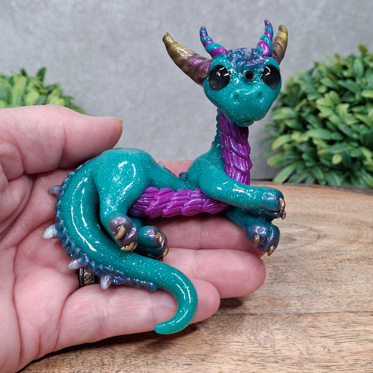  A hand holding a clay dragon against a wood background with greenery