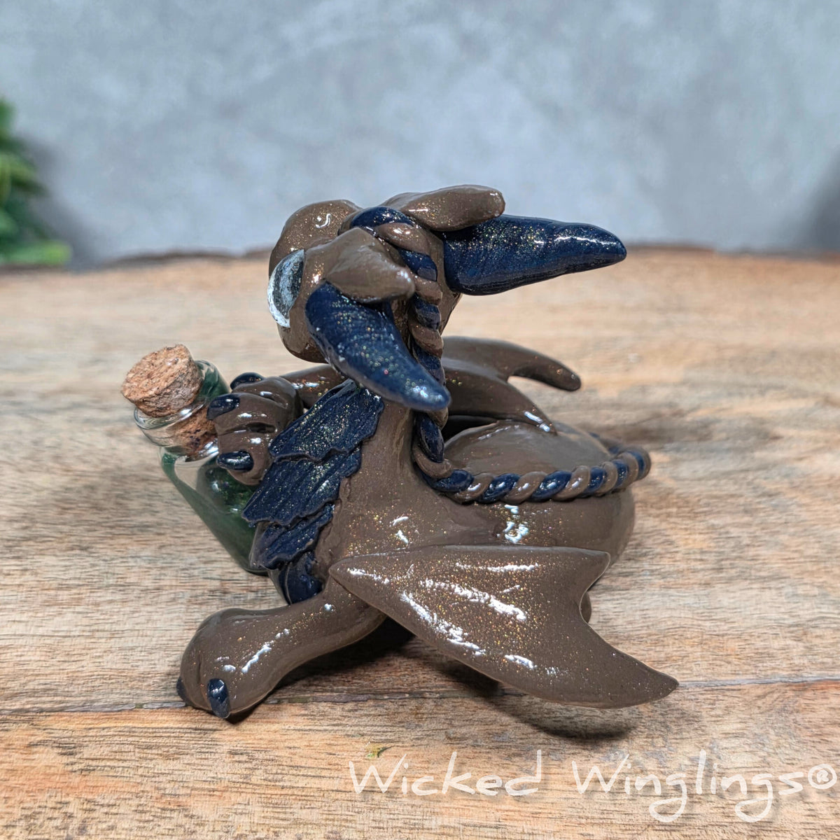 A clay dragon with a potion bottle on a wood surface. Text reads Wicked Winglings.