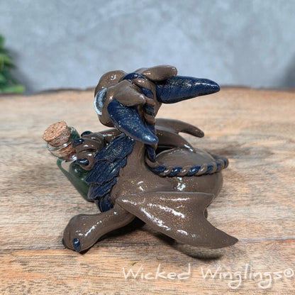 A clay dragon with a potion bottle on a wood surface. Text reads Wicked Winglings.
