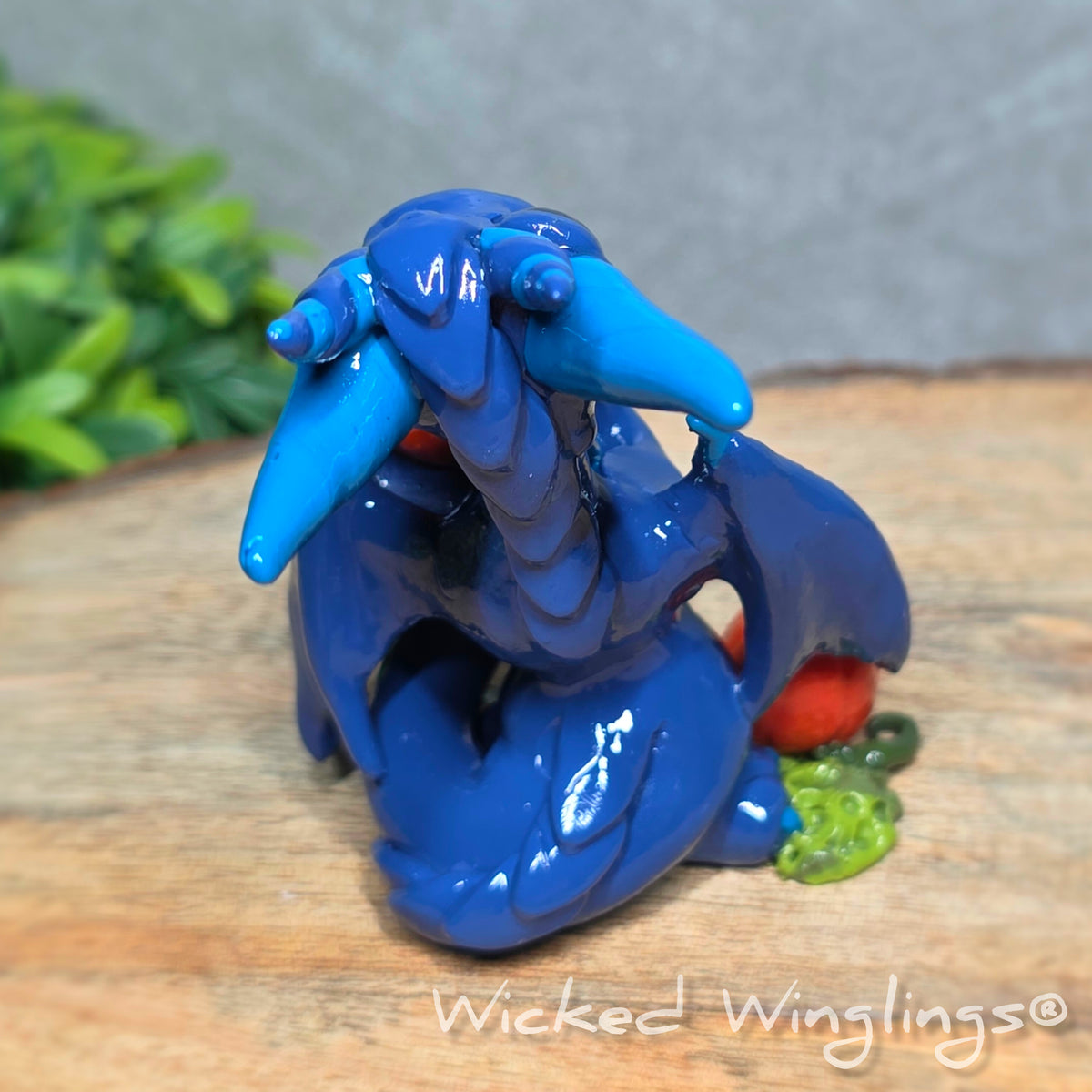Back view of a blue clay dragon with pumpkins against a wood surface with greenery. Text reads Wicked Winglings. 