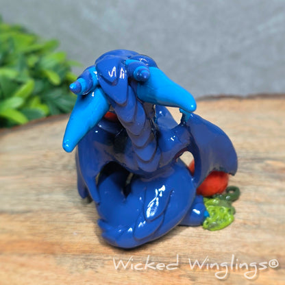 Back view of a blue clay dragon with pumpkins against a wood surface with greenery. Text reads Wicked Winglings. 