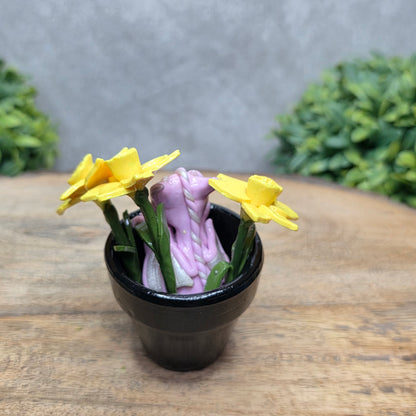 Back view of a clay baby dragon in a pot with daffodils against a wood surface with greenery.