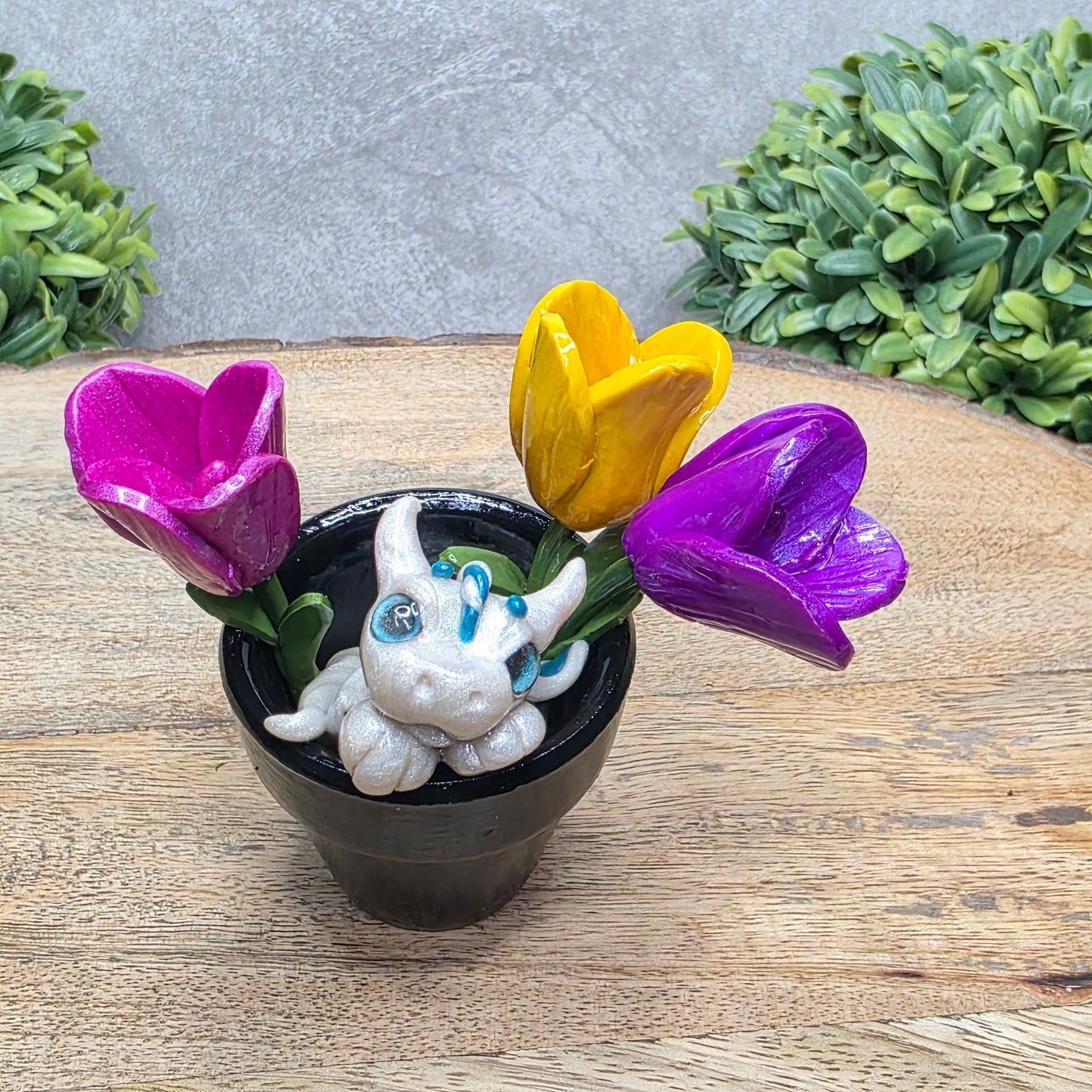 A clay baby dragon in a pot with tulips against a wood surface with greenery.