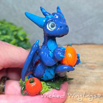 A hand holding a blue clay dragon with pumpkins against a wood surface with greenery. Text reads Wicked Winglings. 
