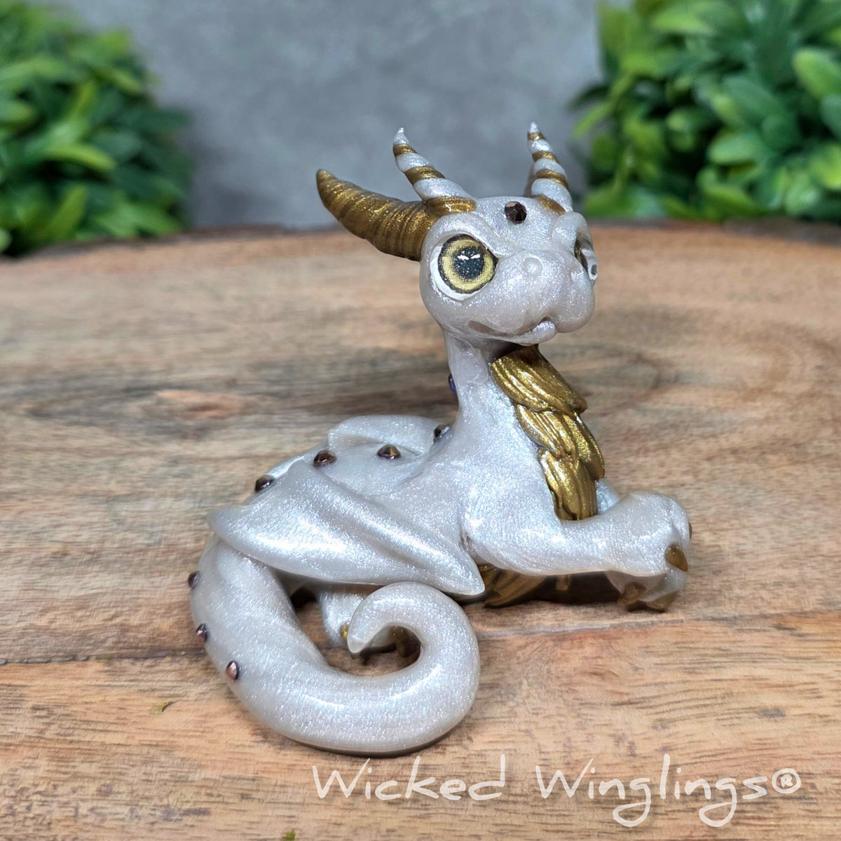 A white clay dragon sitting against a blurred background with greenery. Text says Wicked Winglinng.
